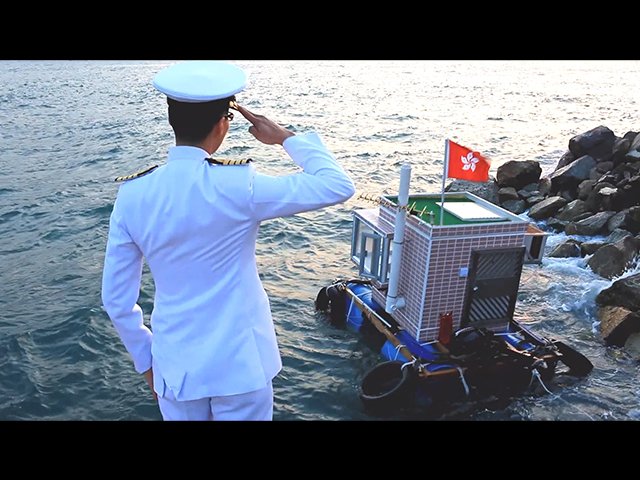Film still showing a man with his back to us in a white captain’s uniform. He salutes a small boat in the water in front of him. The boat consists of a cubic house on a floating platform. It has a door and windows, and a Hong Kong flag on the roof.